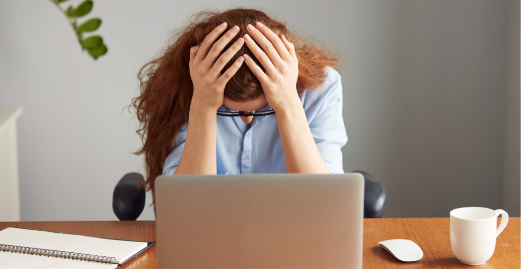 The image portrays a frustrated user, a redheaded woman sitting at her desk with her head in her hands, visibly stressed while looking at her laptop. It effectively represents user frustration, particularly in scenarios where poor interface design or technical issues create negative experiences