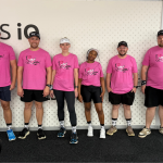 A group of eight Systems iO team members standing in a row inside the office, all wearing matching bright pink breast cancer awareness T-shirts, Systems iO themed socks, and athletic gear for a fun run. They’re smiling and posing in front of a Systems iO branded wall.