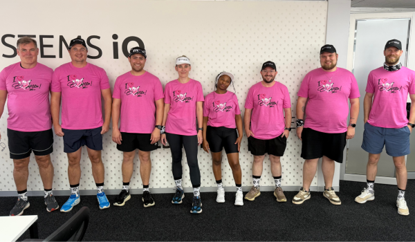 A group of eight Systems iO team members standing in a row inside the office, all wearing matching bright pink breast cancer awareness T-shirts, Systems iO themed socks, and athletic gear for a fun run. They’re smiling and posing in front of a Systems iO branded wall.