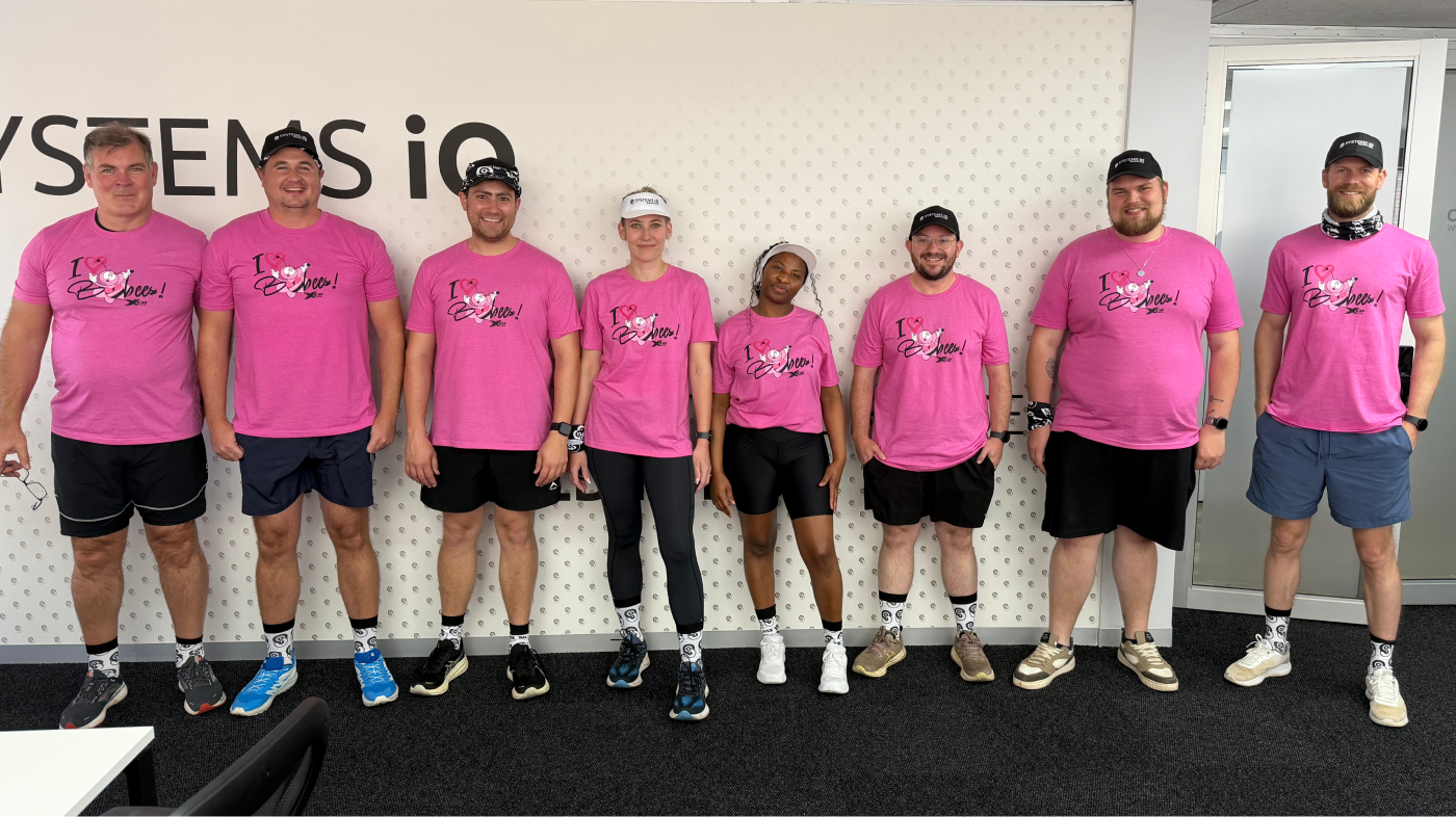 A group of eight Systems iO team members standing in a row inside the office, all wearing matching bright pink breast cancer awareness T-shirts, Systems iO themed socks, and athletic gear for a fun run. They’re smiling and posing in front of a Systems iO branded wall.